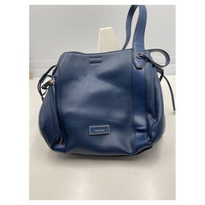 May include: A navy blue leather handbag with a shoulder strap and a zipper pocket. The bag has a textured surface and a small silver Calvin Klein logo. The bag is sitting on a white surface.