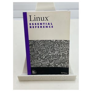 May include: A book titled "Linux Essential Reference" is displayed on a white stand. The book has a white cover with the title in large, dark blue letters and a purple spine. The lower half of the cover features a black and white image.