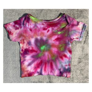 May include: A tie-dye baby bodysuit in shades of pink, purple, and green. The short-sleeved shirt has a round neckline and is made of soft fabric. The colorful pattern is centered on the front of the shirt.