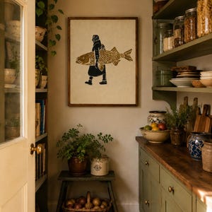 May include: A framed wall art print featuring a block-print style illustration of a person in a headscarf carrying a large golden-brown fish. The artwork is displayed on a neutral wall in a kitchen setting with wooden shelves and cabinets.