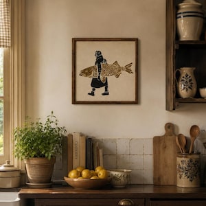 May include: A square framed wall art piece featuring a black and gold linocut-style print of a person carrying a large fish. The artwork is displayed on a light-colored wall above a kitchen counter, showcasing a detailed graphic illustration.