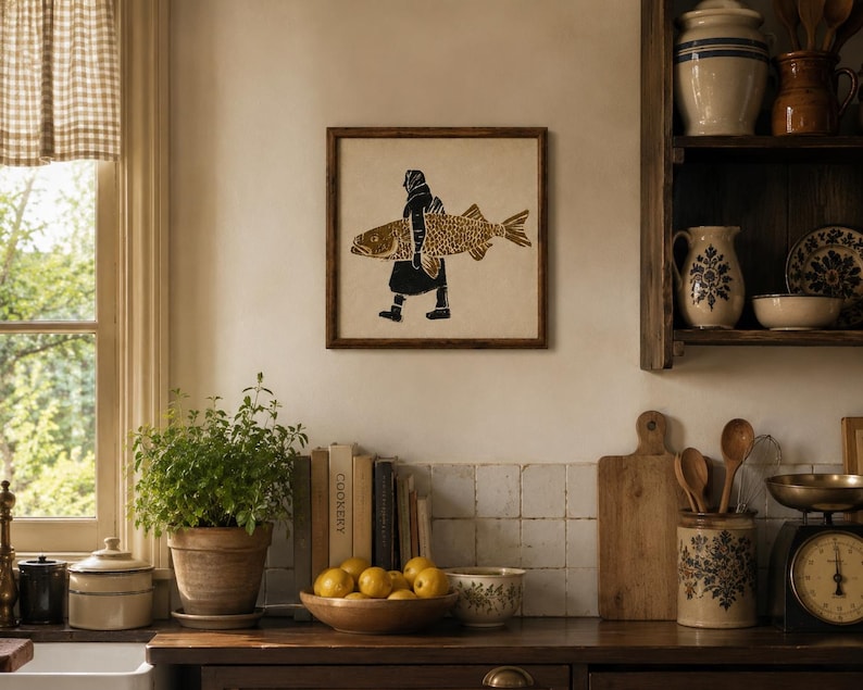 May include: A square framed wall art piece featuring a black and gold linocut-style print of a person carrying a large fish. The artwork is displayed on a light-colored wall above a kitchen counter, showcasing a detailed graphic illustration.