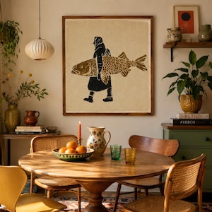 May include: A square framed wall art print featuring a black and gold linocut-style illustration of a person carrying a large fish. The artwork is displayed on a light-colored wall above a round wooden dining table with chairs and decor.