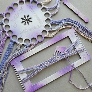Puede incluir: Un organizador de hilos circular con degradado morado y blanco con agujeros numerados, un telar rectangular con grabados florales, un pequeño peine a juego y una pulsera con patrón de diamantes parcialmente tejida con hilo de bordar morado y crema.