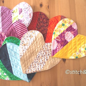 May include: A selection of heart-shaped quilted items in diverse colours and patterns, including floral, striped, and solid fabrics. The hearts are arranged on a wooden surface, showcasing a handmade craft theme.