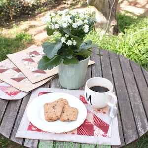 Könnte beinhalten: Ein runder Holztisch, der für einen entspannten Moment im Freien gedeckt ist. Er zeigt eine weiße Tasse mit Kaffee, einen Teller mit Keksen und eine Topfpflanze mit weißen Blüten. Gesteppte Tischsets verleihen der Szene einen Hauch von Farbe und Muster.