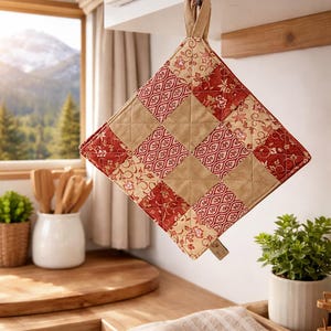 May include: A quilted square pot holder with a loop for hanging. The pot holder features a patchwork design with alternating squares of beige and red floral and patterned fabric. The kitchen accessory is hanging in a kitchen setting.