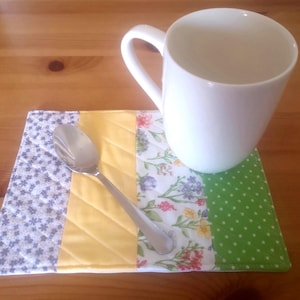 May include: A white ceramic mug sits on a patchwork placemat with floral, yellow, and green polka dot sections. A silver spoon rests on the placemat. The placemat is on a wooden table.