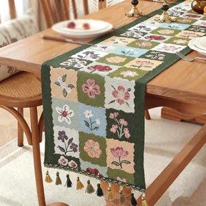 May include: A floral table runner with a dark green border and a patchwork design featuring various flowers in pink, white, and purple. The runner is adorned with small tassels along the bottom edge. The runner is placed on a wooden table.