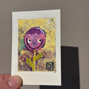 May include: A small, whimsical painting of a flower with a smiling face. The flower has a purple head with dark spots, a yellow stem, and green leaves. The background is gold with a white border.
