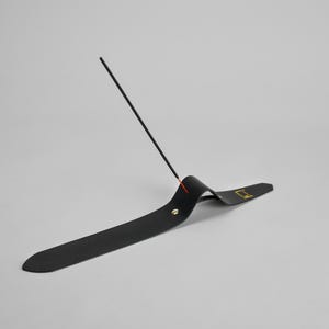 Puede incluir: Un porta incienso negro con un diseño curvo, con un logotipo dorado y un pequeño acento rojo. Un palito de incienso se coloca en el soporte, creando una estética minimalista.