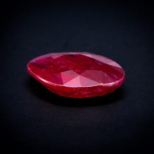 May include: A faceted oval-shaped gemstone with a deep red color and a polished surface. The stone displays multiple geometric cuts that reflect light across its translucent body, showcasing a vibrant crimson hue against a dark background.