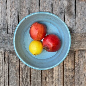 Può includere: Una ciotola in ceramica rotonda di colore azzurro con finitura maculata contenente tre frutti: un limone giallo brillante, una nettarina rossa liscia e un melograno rosso strutturato, disposti insieme al centro della ciotola.