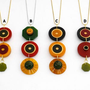 May include: Four handmade necklaces with gold chains. Each necklace features a unique design of stacked, colorful circular and geometric shapes. Colors include red, orange, black, green, and yellow. The necklaces are displayed against a white background.