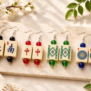 May include: Four pairs of earrings featuring ivory-colored Mahjong tiles with various symbols and numbers. Each earring has a different color bead: black, red, green, and blue. The earrings are displayed on a light-colored surface with floral accents.