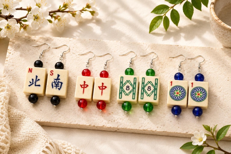 May include: Four pairs of earrings featuring ivory-colored Mahjong tiles with various symbols and numbers. Each earring has a different color bead: black, red, green, and blue. The earrings are displayed on a light-colored surface with floral accents.