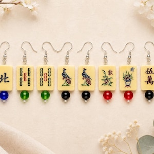 May include: A collection of eight earrings featuring rectangular cream-colored tiles with various designs and Chinese characters. Each earring has a silver hook and a colorful bead, including blue, green, black, and red. The earrings are displayed on a light beige surface.