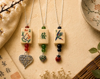 Puede incluir: Tres collares de cadena plateada con colgantes de fichas de mahjong. El primero tiene una ficha floral, cuentas rojas y un corazón. El segundo tiene una ficha de carácter verde, cuentas verdes y un corazón pequeño. El tercero tiene una ficha de pájaro, cuentas negras y una mariquita.