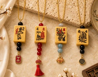 Puede incluir: Cuatro collares dorados con colgantes que se asemejan a fichas de Mahjong. Cada colgante es de color crema con ilustraciones y números en negro y rojo. Los collares tienen cadenas doradas, cuentas negras y amuletos decorativos con caracteres chinos. El fondo es beige claro.
