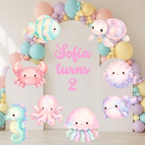 Puede incluir: Un fondo de fiesta con un panel arqueado blanco con el texto rosa Sofia turns 2, rodeado por un arco de globos en tonos pastel. Las criaturas marinas en acuarela incluyen un pez, tortuga, cangrejo, pez globo, caballito de mar, pulpo, ballena y medusa.