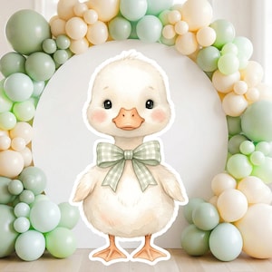 May include: A charming illustration of a duckling with a green plaid bow tie. The duckling has a white body, orange beak, and rosy cheeks. The background features a white circle and a balloon arch in shades of green and cream.