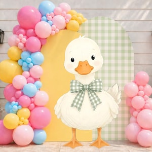 Puede incluir: Ilustración de un pato blanco con una pajarita a cuadros verdes, sobre un fondo de globos pastel y un arco amarillo. La escena está decorada con un patrón de cuadros verdes y blancos, creando un ambiente alegre y festivo.