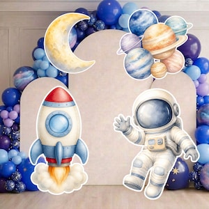 May include: A space-themed party decoration set featuring a large yellow crescent moon, a cluster of colorful planets, a red and white rocket ship with orange exhaust, and an astronaut in a white suit, all set against a blue balloon arch.