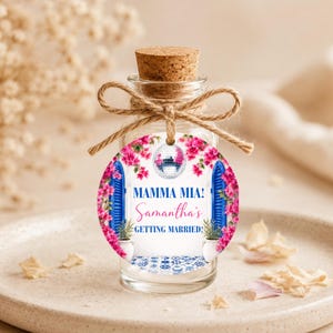 Może przedstawiać: Mała szklana butelka z korkiem i ozdobną etykietą. Etykieta zawiera napis "MAMMA MIA! Samantha's GETTING MARRIED!" z kulą dyskotekową, niebieskimi okiennicami i różowymi kwiatami. Butelka jest związana sznurkiem.