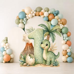 May include: A decorative cutout featuring a large number one with a green scale pattern, a small green dinosaur, a volcano, and a cracked egg. The scene is surrounded by a large arch of balloons in shades of blue, green, tan, and white.