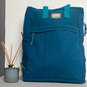May include: Teal tote bag with teal handles and a front zipped pocket. The bag features a small patch with text and a floral design. A small ceramic diffuser with reeds is in the foreground.