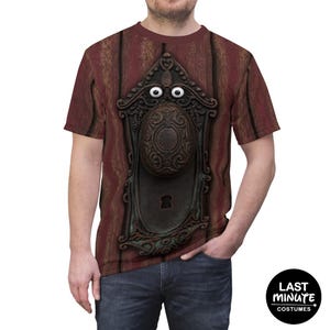 May include: A short-sleeve t-shirt featuring a graphic print of a dark brown, ornate, vintage-style door handle and keyhole plate set against a vertical wood-grain background. Two white, round, cartoon-style eyes are positioned above the knob.