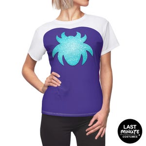 Puede incluir: Camiseta blanca y morada con un diseño brillante turquesa en la parte delantera. La camiseta tiene mangas cortas y cuello redondo. El texto "LAST MINUTE COSTUMES" está impreso en un círculo negro.