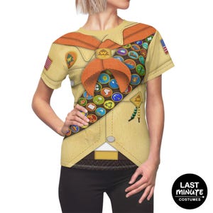 May include: A tan t-shirt featuring a printed scout uniform design. The design includes a brown neckerchief, merit badges, and a gold-colored emblem. The shirt has short sleeves and a printed American flag on the sleeve.