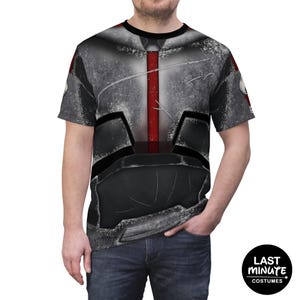 May include: A gray and black t-shirt featuring a detailed armor design with a red vertical stripe. The shirt has a crew neck and short sleeves. The design is printed on the front of the shirt. The shirt is worn by a person.