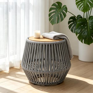 May include: A round side table featuring a light wood top and a base constructed from thick, vertical grey rope strands. A white candle in a glass jar and a folded grey knit textile are placed on the flat wooden surface of the table.