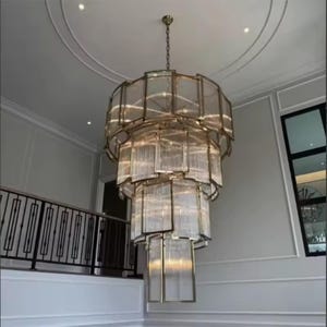 May include: A large, multi-tiered chandelier with a gold finish and rectangular glass panels. It hangs from a white ceiling. The chandelier's design is layered, creating a cascading effect. It is suspended by a chain.