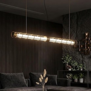 Könnte beinhalten: Eine moderne, lineare Pendelleuchte mit warmem, einladendem Licht. Die Leuchte hat ein zylindrisches Design mit einem goldfarbenen Metallrahmen und einer hellen, weißen Lichtquelle. Die Leuchte hängt an dünnen Drähten und erzeugt eine minimalistische Ästhetik.