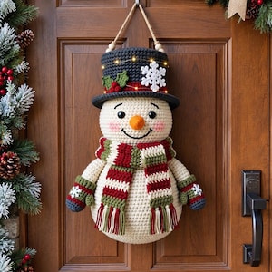 May include: A crocheted snowman wall hanging featuring a dark gray top hat with a white snowflake and holly berries, a striped red, green, and cream scarf, and matching mittens. The snowman has a bright orange nose and small LED string lights.