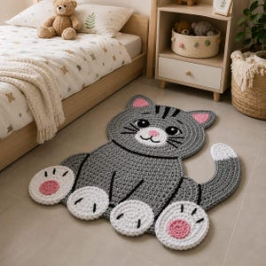 May include: A gray crocheted cat-shaped rug featuring a smiling face with black eyes, a pink nose, and pink inner ears. The rug has four white paws with pink circular pads and a curved tail with a white tip, all crafted in a thick yarn texture.