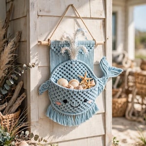 May include: A light blue macrame wall hanging shaped like a whale with a pocket containing seashells and two starfish. The whale features a white fringe spout, black embroidered eyes, a small smile, and pink cheeks, suspended from a wooden dowel.