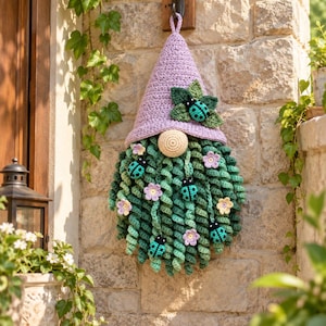 May include: A crochet gnome wall hanging featuring a tall lavender pointed hat adorned with green leaves and a turquoise ladybug. The gnome has a round beige nose and a long, curly dark green beard decorated with small flowers and ladybugs.