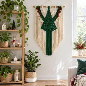 Könnte beinhalten: Ein Makramee-Wandbehang mit einem zentralen, dunkelgrünen Weihnachtsbaum-Design aus geknüpfter Kordel, verziert mit kleinen roten und goldenen Ornamenten, Tannenzapfen und langen cremefarbenen Fransen, aufgehängt an einem Holzstab.