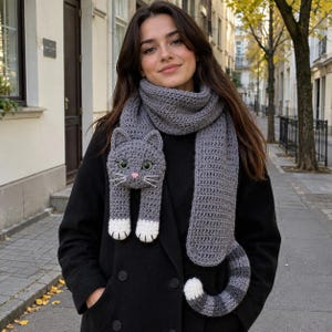 May include: A gray crocheted scarf featuring a cat design with a head, white-tipped paws, and a striped tail. The cat head has green eyes, a pink nose, and white whiskers. The scarf is made of a textured yarn and is worn over a black coat.