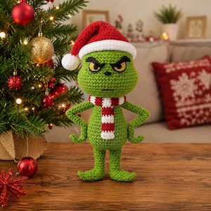 Op de afbeelding: Een groene gehaakte pop met een norse gezichtsuitdrukking, gekleed in een rood-witte kerstmuts en een bijpassende rood-wit gestreepte sjaal. De pop heeft een rond hoofd, puntige voeten en staat met de handen in de zij op een houten ondergrond.