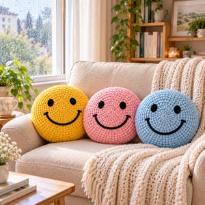 May include: Three round, crocheted pillows in yellow, pink, and light blue, each featuring a black embroidered smiley face with two circular eyes and a curved mouth. The pillows are arranged side-by-side on a textured, light-colored sofa.