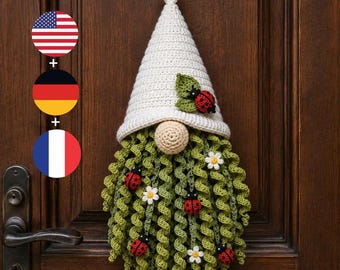 May include: A crochet gnome wall hanging featuring a tall, off-white pointed hat adorned with a green leaf and a red ladybug. The gnome has a round beige nose and a long, curly green beard decorated with small white flowers and red ladybugs.