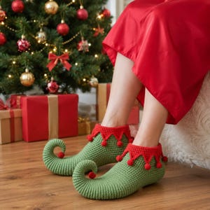 Peut inclure: Une paire de chaussons en crochet vert avec des bouts recourbés et des pompons rouges le long du bord supérieur. Les chaussons présentent un revers rouge avec un motif festonné et de petits pompons rouges attachés à chaque pointe.