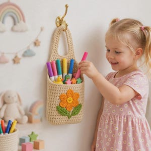 May include: A beige crochet hanging basket featuring a large orange flower with green leaves on the front. The basket is suspended from a gold-colored metal wall hook and contains several colorful markers and small plastic animal figurines.