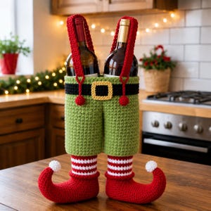 May include: A crochet wine bottle holder designed as elf pants with green trousers, a black belt with a yellow buckle, red suspenders, and red curled-toe shoes with white pom-poms. The legs feature red and white horizontal stripes above the feet.