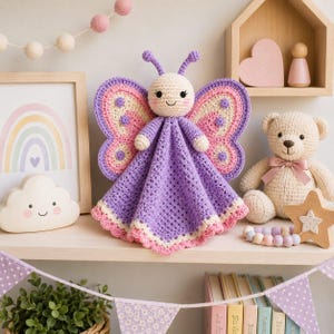 May include: A crochet butterfly lovey featuring a cream-colored head with embroidered facial features, purple antennae, and large wings in shades of pink, cream, and purple. The body consists of a purple textured blanket with a scalloped pink edge.